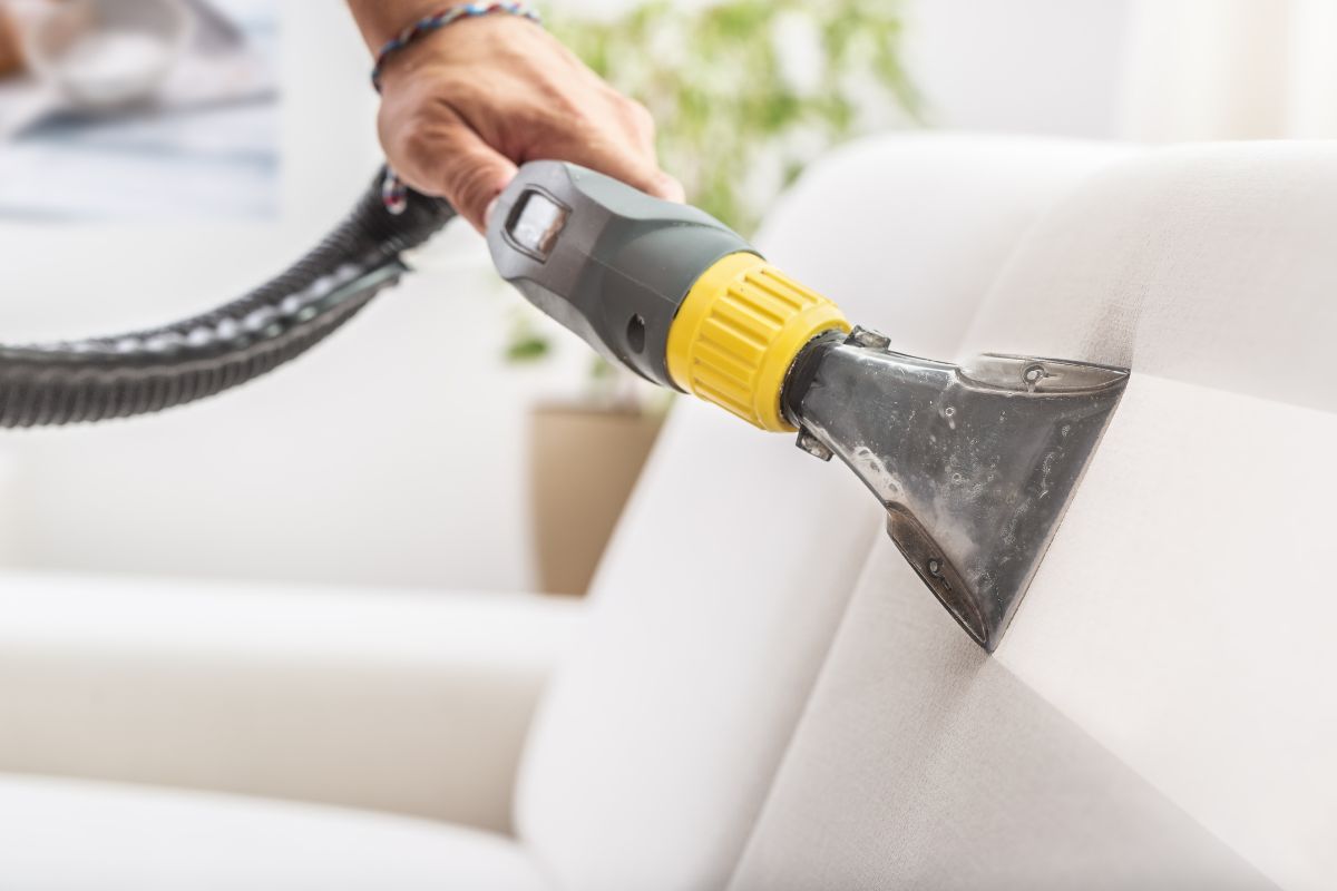 A person using a vacuum to clean the seat of a couch.